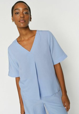 Nő világoskék V-nyakú, rövid ujjú felsőt és hozzáillő nadrágot visel, és egy egyszerű fehér háttér előtt áll.