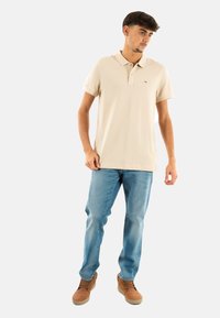 Beige polo tričko s krátkým rukávem, klasickým límečkem a vyšitým logem. Nosené s světle modrými džíny a hnědými šněrovacími botami.