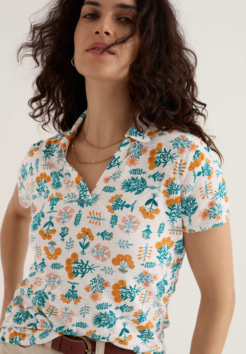 Femme aux cheveux bouclés et foncés portant une chemise blanche à manches courtes avec des motifs floraux orange et verts et deux colliers en or.