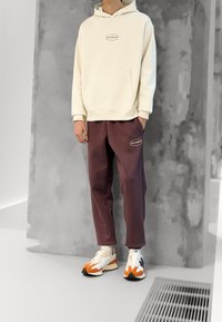 Felpa beige con tasca a marsupio, pantaloni della tuta viola con patch logo e sneakers multicolori con accenti arancioni e blu.