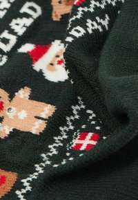 Dzianinowy kocyk o ciemnozielonym tle, ozdobiony wzorami piernikowych ludzików, twarzy Świętego Mikołaja oraz płatków śniegu w beżowych, czerwonych i białych kolorach. Miękka faktura.