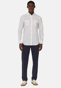 White long-sleeve shirt with two breast pockets, gold buttons, paired with dark navy pants and white sneakers with beige soles.