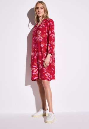 Rotes Blumen-Kleid mit langen Ärmeln, V-Ausschnitt und elastischen Bündchen, aus leichtem Stoff, kombiniert mit weißen Sneakers.