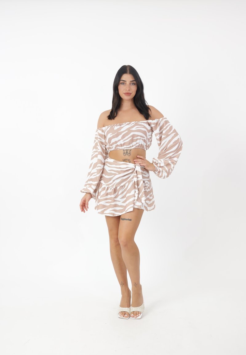 Frau steht, trägt ein beiges und weißes Zebra-Off-Shoulder-Top und einen passenden Rock mit offenen Zehenabsätzen vor einem schlichten weißen Hintergrund.