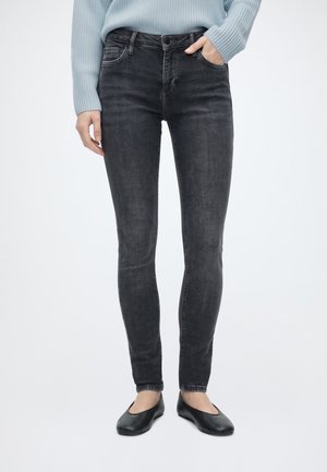 Eine Frau trägt ausgewaschene schwarze Skinny-Jeans, einen hellblauen Pullover und schwarze Ballerinas und steht mit einer Hand in der Tasche.