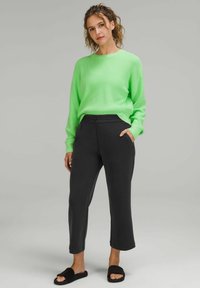 Neonowa zielona swetrowa bluzka z prążkami i czarne rybaczki z bocznymi kieszeniami. Modelka ma na sobie czarne sandały na płaskiej podeszwie, a jej fryzura jest falista.