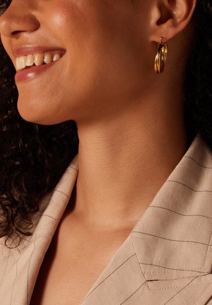 Gouden hoopoorbellen met een gepolijste afwerking, met onderling verbonden designelementen. Het model draagt een lichtbeige gestreepte blazer.