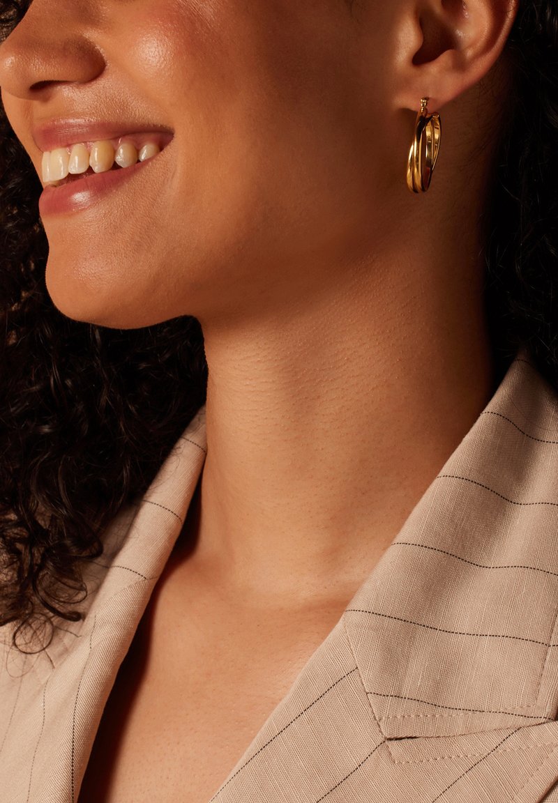 Gouden hoopoorbellen met een gepolijste afwerking, met onderling verbonden designelementen. Het model draagt een lichtbeige gestreepte blazer.
