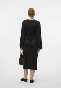 Abito nero a coste con maniche lunghe e vita legata. La modella tiene una borsa marrone e indossa delle ballerine nere, vista da dietro su uno sfondo bianco.