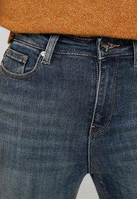 Jeans de mezclilla azul oscuro con un diseño clásico de cinco bolsillos, que cuentan con un cierre de cremallera y costuras en contraste a lo largo de la cinturilla y las costuras.