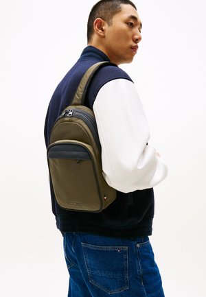 Olive green backpack made of smooth, durable material with a front zip compartment, worn on one shoulder against a navy and white jacket.