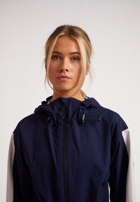 Chaqueta con capucha de color azul marino que presenta un frontal con cremallera, cordón ajustable y acentos blancos en los hombros y las mangas. Tejido suave y ligero.