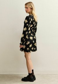Robe noire à fleurs avec des manches longues, une taille cintrée et une jupe évasée. Présente des fleurs roses et blanches avec des accents verts. Portée avec des chaussures noires.