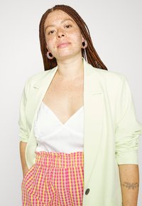 Blazer verde claro sobre um top de seda branca, combinado com calças de cintura alta em padrão xadrez rosa e amarelo. Cabelo trançado notável e brincos circulares.