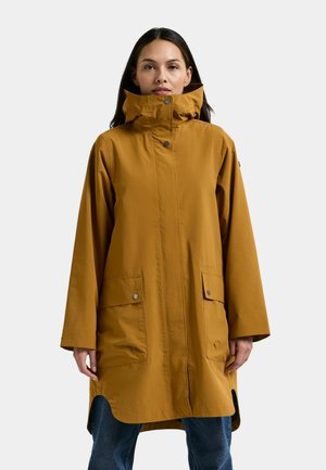 Kvinde iført en lang sennepsgul regnfrakke med høj krave og frontlommer med klap, kombineret med blå jeans mod en ensfarvet baggrund.