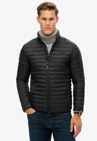 Chaqueta de plumas negra con diseño de acolchado horizontal, cierre con cremallera, dos bolsillos laterales y cuello alto, llevada sobre un cuello de tortuga gris.