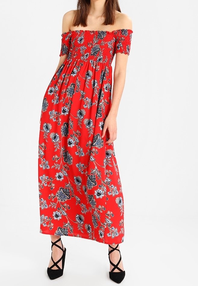 Femme portant une robe longue rouge épaules dénudées avec des motifs floraux bleus et blancs et des talons noirs à brides croisées autour de la cheville.