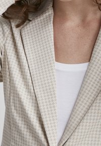 Béžový vzorovaný blazer s texturovanou, tkanou látkou a klopovým límcem, položený na bílém topu. Viditelné švy podél okrajů.