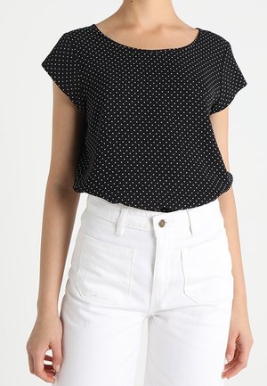 Femme portant un haut noir à manches courtes avec des pois blancs rentré dans un pantalon blanc taille haute avec poches avant et fermeture à boutons.