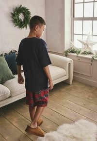 Marineblaues Oversize-T-Shirt, rote Karo-Shorts und braune Hausschuhe an einem Kind, in einem lichtdurchfluteten Raum mit einer weißen sternförmigen Dekoration und Pflanzen.