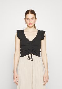 Top de tanque preto com nervuras, decote em V, centro franzido e mangas com babados; combinado com uma saia plissada bege.