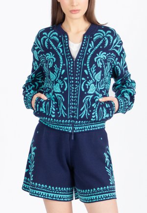 Mujer con chaqueta de punto azul marino con cremallera y pantalones cortos a juego con patrones de palmeras tropicales en azul claro, con las manos en los bolsillos de la chaqueta.