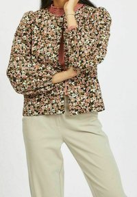 Kvinna iklädd blommig mönstrad långärmad blus med puffärmar och beige hög midja byxor, hand vilande på ansiktet och armarna korsade.