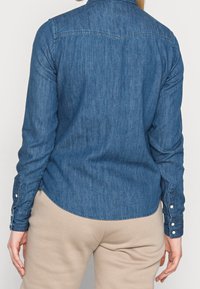 Denim shirt in dark blue, features a classic collar and buttoned cuffs, with a slightly curved hem and textured fabric.