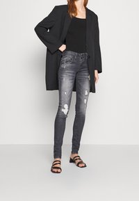Blazer negro sobre un top negro sin mangas, combinado con jeans gris desgastados y sandalias de tacón negro con tiras. Los jeans presentan detalles rasgados.