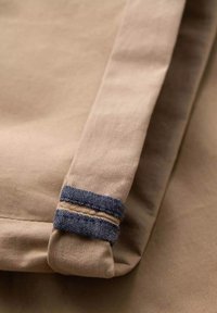 Tissu en coton beige avec une texture lisse, présentant un poignet avec des rayures bleu marine et des détails de couture.