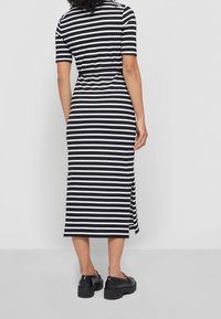 Nainen, jolla on yllään mustavalkoinen vaakasuora raidallinen midi-mekko lyhyillä hihoilla ja mustat platform-loafers-kengät, seisoo yksinkertaisella taustalla.