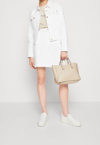 Donna che indossa una giacca bianca, una gonna bianca, una t-shirt beige e delle scarpe slip-on bianche, mentre tiene una borsa beige contro uno sfondo bianco semplice.