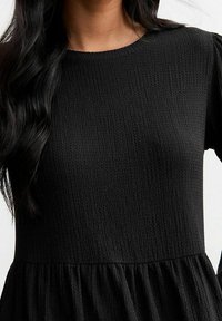 Schwarzes Kleid aus strukturiertem Stoff mit rundem Ausschnitt und locker sitzenden Ärmeln, die in einem gerafften, ausgestellten Saum enden.