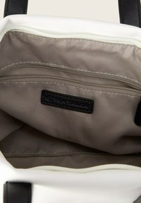 L'intérieur du sac fourre-tout blanc présente un revêtement gris clair, une poche zippée et une étiquette en cuir noir avec "TOM TAILOR" embossé.