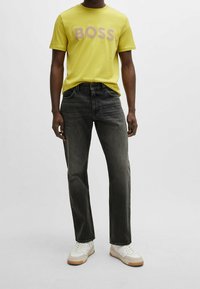 Camiseta de algodón amarilla con el logo "BOSS" en rosa, combinada con vaqueros gris oscuro y zapatillas blancas con suelas beige.