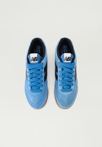 Adidași din piele întoarsă albastră cu dungi negre pe laterale și șireturi albastre. Talpă din cauciuc, vârf rotunjit și etichetă cu logo pe limbă. Design simplu.