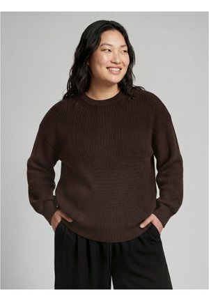 Femme aux longs cheveux foncés portant un pull en tricot marron foncé et un pantalon noir, souriante et regardant sur le côté, les mains dans les poches.