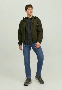 Giacca a vento verde scuro con chiusura a zip, indossata sopra una camicia a quadri grigia, abbinata a jeans blu e sneaker grigie.