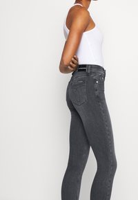 Jeans skinny negros de denim, con una cintura alta, bolsillos traseros con detalles de costura y un logo visible en la cinturilla.