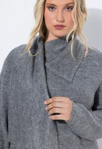 Grijze gebreide trui met een hoge kraag en een asymmetrisch ontwerp. De structuur is geribbeld, wat een gezellige, gelaagde uitstraling geeft.
