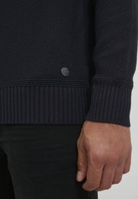 Mörkblå stickad tröja med texturerad kropp, ribbade ärmslut och nederkant. Har en liten rund logobadge på nedre vänstra sidan.