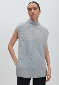 Frau mit nach hinten gekämmtem Haar, die einen ärmellosen, grauen, hochgeschlossen gestrickten Pullover und schwarze Hosen trägt, vor einem einfarbigen Hintergrund.