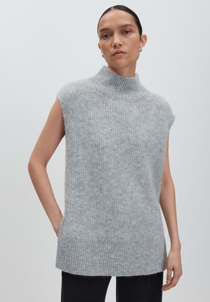 Donna con i capelli all'indietro che indossa un gilet di maglina grigia a collo alto senza maniche e pantaloni neri, su uno sfondo semplice.