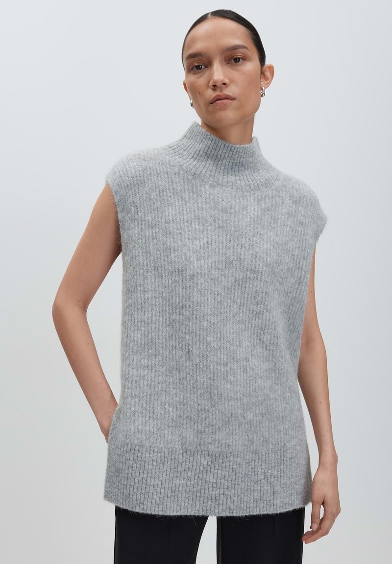 Frau mit nach hinten gekämmtem Haar, die einen ärmellosen, grauen, hochgeschlossen gestrickten Pullover und schwarze Hosen trägt, vor einem einfarbigen Hintergrund.