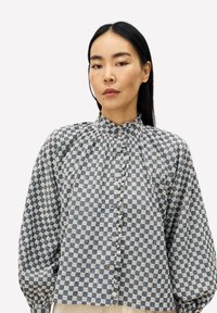 Frau mit langen schwarzen Haaren trägt eine hochgeschlossene blau-weiße karierten Bluse mit Frontknöpfen und puffigen langen Ärmeln.