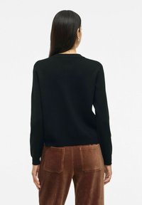 Pull en maille noir à manches longues et col rond, présenté de dos. Associé à un pantalon en velours côtelé marron avec de grandes poches.
