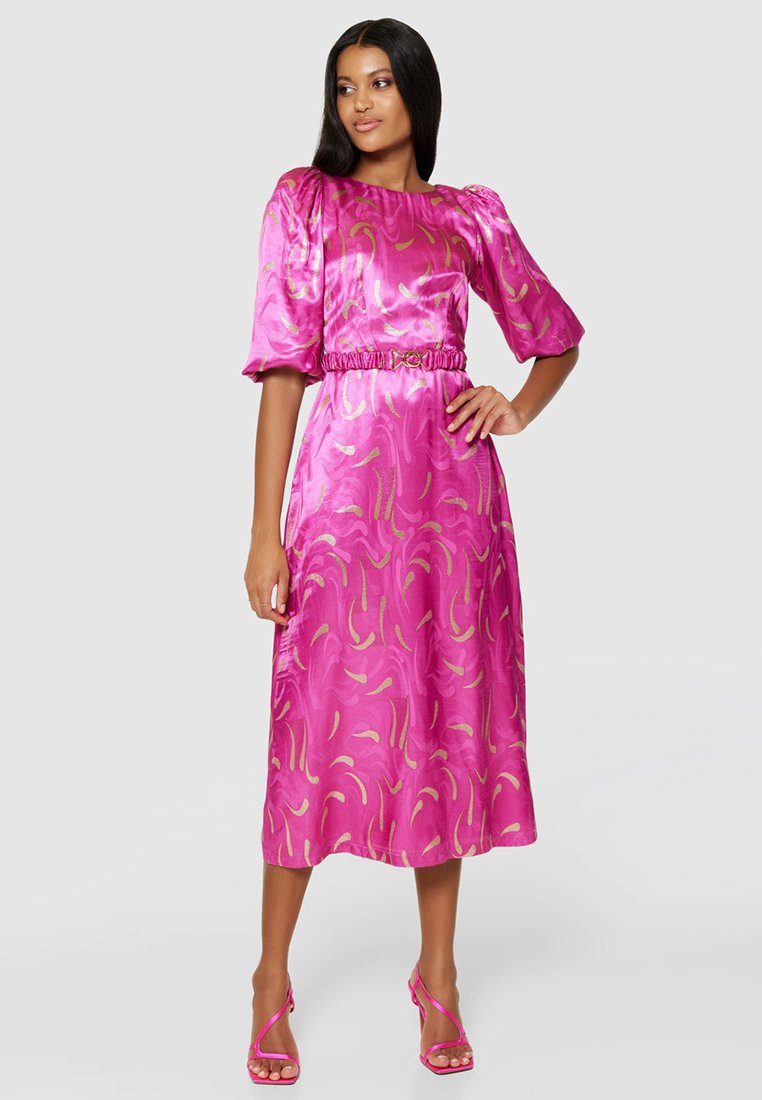 Donna che indossa un elegante abito midi in satin rosa brillante con motivi dorati, maniche a sbuffo, vita cintata e sandali con tacco alto rosa abbinati.