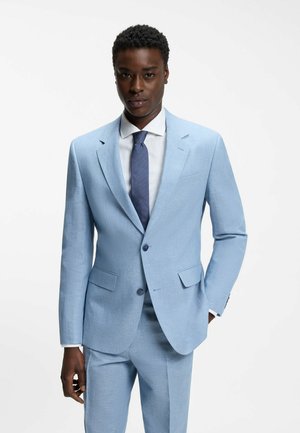 Homme portant un costume bleu clair avec une chemise blanche et une cravate bleu foncé, debout avec une main dans la poche devant un fond uni.