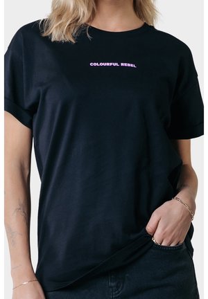 Kobieta w czarnym T-shircie z różowym napisem "COLOURFUL REBEL" na piersi, ręka oparta w kieszeni ciemnych dżinsów, tatuaże na lewym ramieniu.