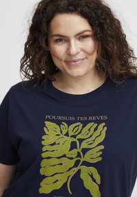 Tamsiai mėlynos medvilninės marškinėlių su žaliu lapo grafiku, turinčiu iškirptus detales, ir tekstu "POURSUIS TES REVES" virš dizaino.